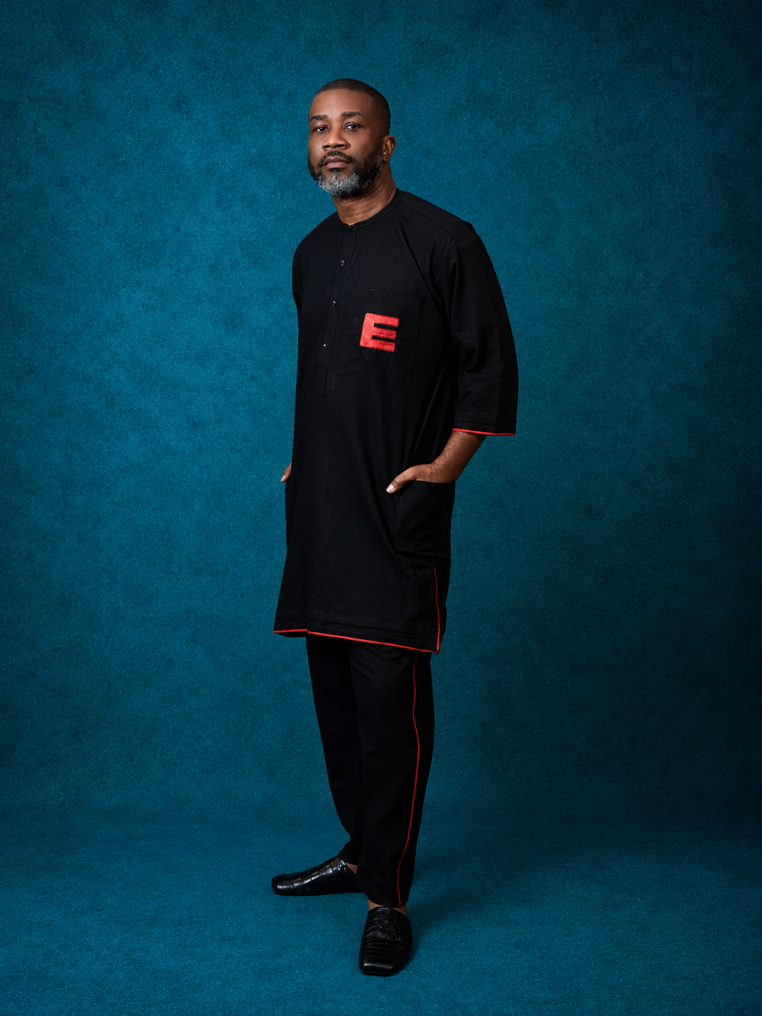 Model wearing an OBIDA black kaftan with red trim and embroidered detail against a teal studio backdrop.